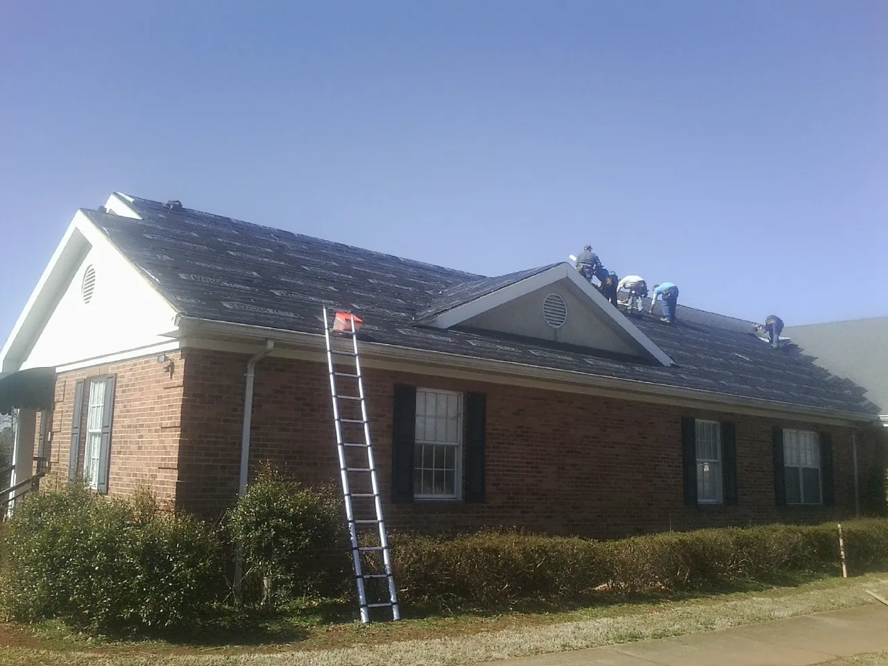 Crew tearing shingles off a brick ranch roof with a ladder set — Emergency Roof Repair work by our crew in Godley