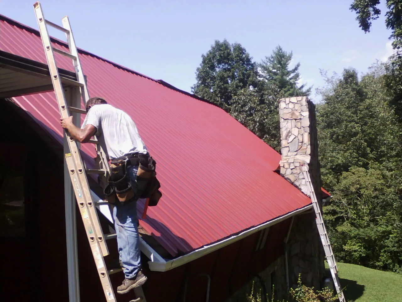 Roofer on a ladder beside a red metal roof and stone chimney — Soffit & Fascia Repair by Roofing Brothers in Godley